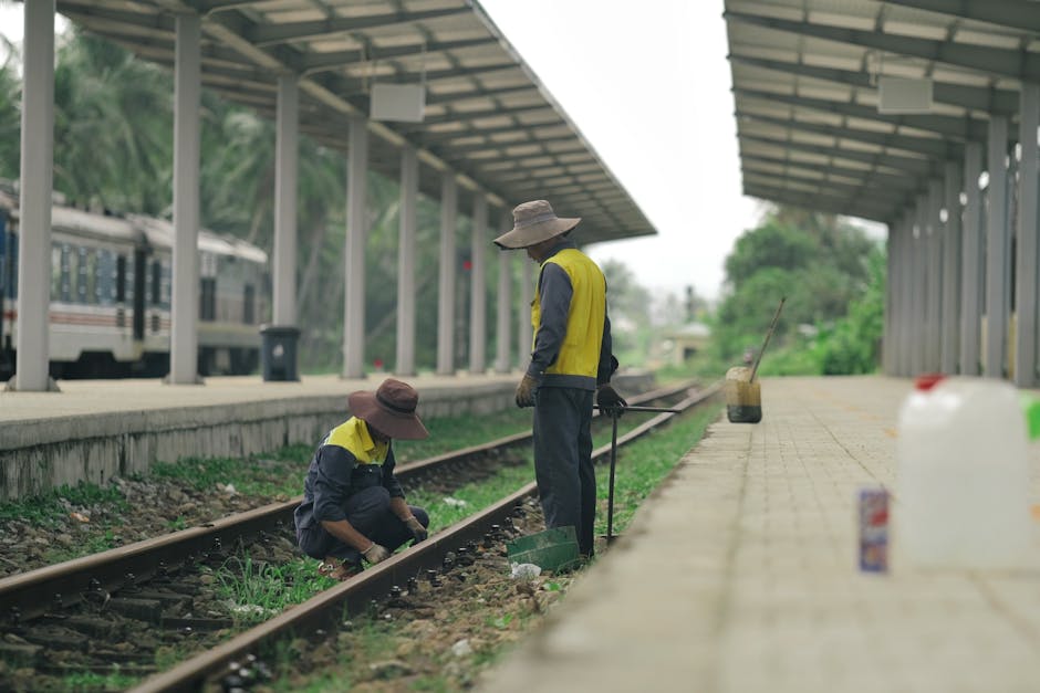 Two railway workers in hats maintain train tracks at a rural station.